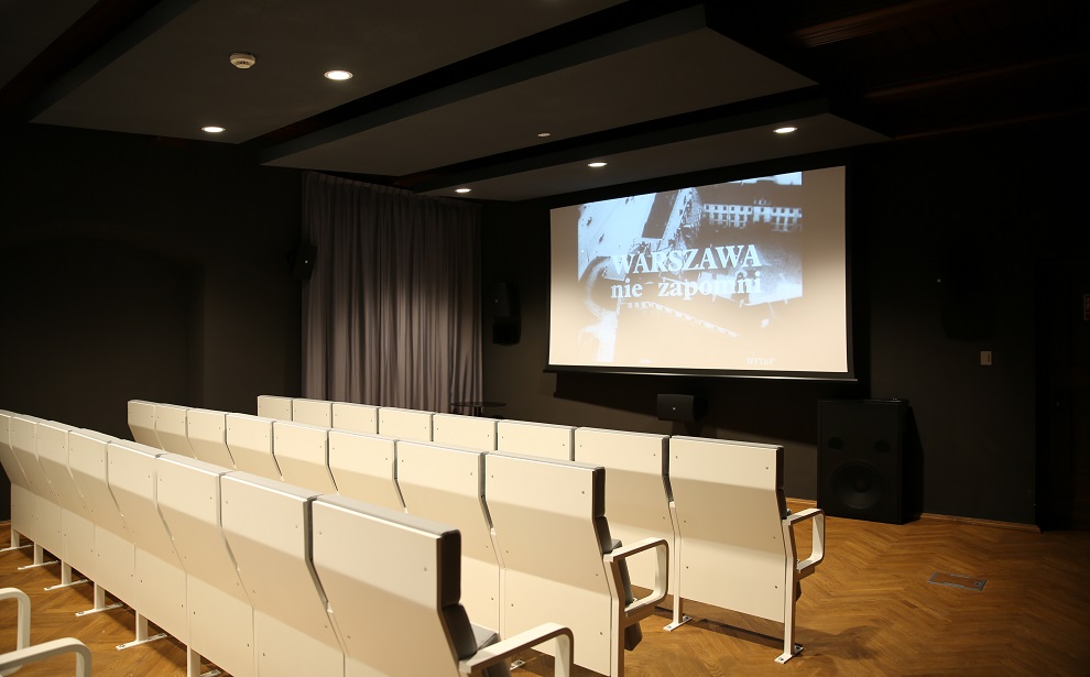 Powiększ obraz: Na zdjęciu: Wnętrze sali kinowej. Na pierwszym planie rzędy białych pleców foteli, w tle ekran z początkowym kadrem z filmu.